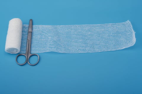 Close-up of surgical scissors and a roll of sterile gauze with an extended strip, placed on a blue background