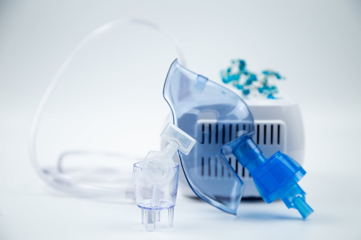 Close-up of an inhalation bowl with an ampoule of medicinal solution in it against the background of a blue breathing mask and a device on a white background.