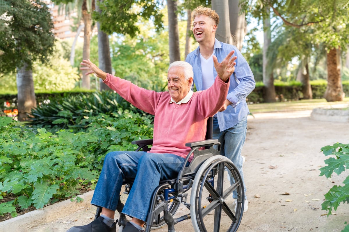 Happy grandson pushing smiling grandfather in wheelchair in park