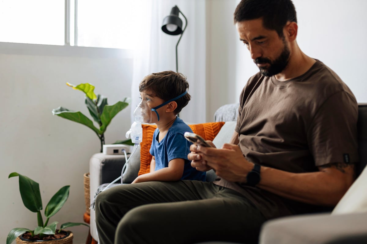 Father sitting beside child during nebulizer treatment at home, illustrating emotional support, pediatric healthcare and daily family caregiving routine.