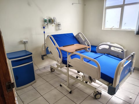 Hospital room with two blue and beige patient beds equipped with IV stands, beside a rolling storage cabinet on a tiled floor