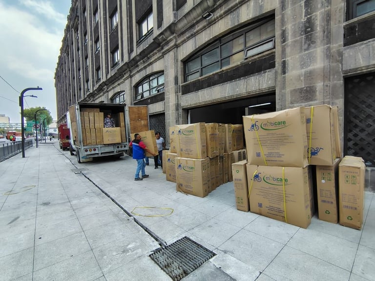 Moving truck and stacked cardboard boxes on urban street next to large building, worker in blue jacket nearby