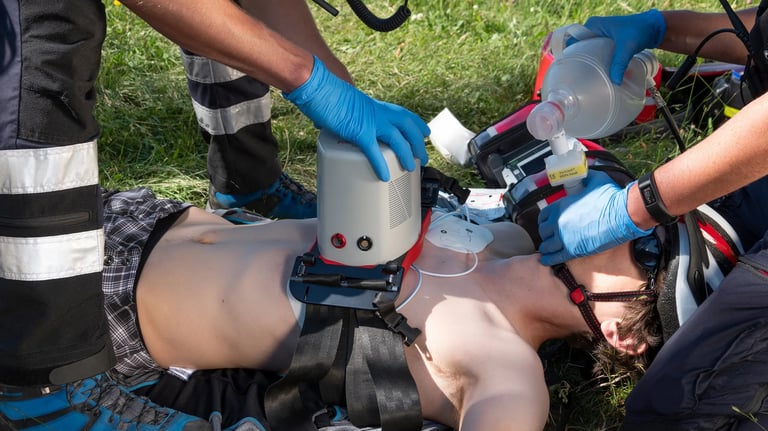 Two people wearing blue gloves operating emergency medical equipment outdoors on grass during what appears to be a training or rescue operation